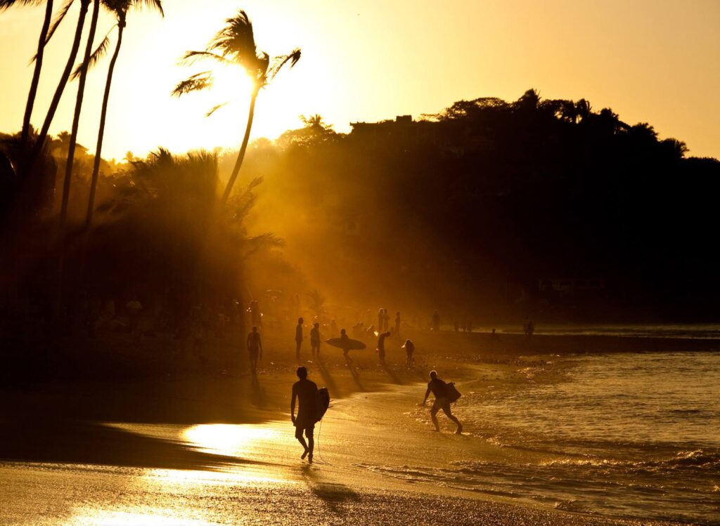 Atardecer en playa con surfistas, palmeras y mar.